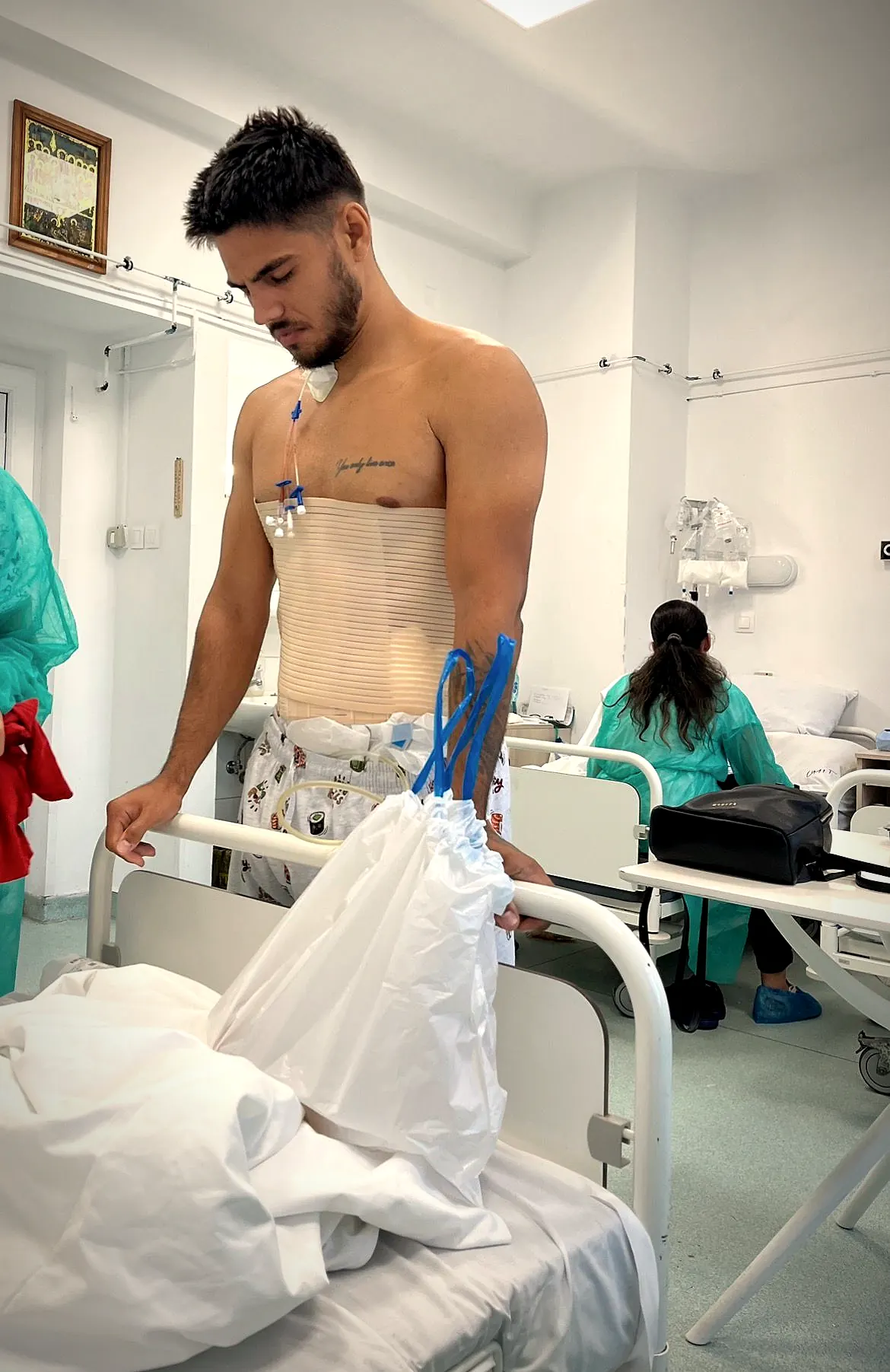Cătălin în salonul de spital după operație, cu brâu abdominal și cateter, sprijinit de marginea patului.
